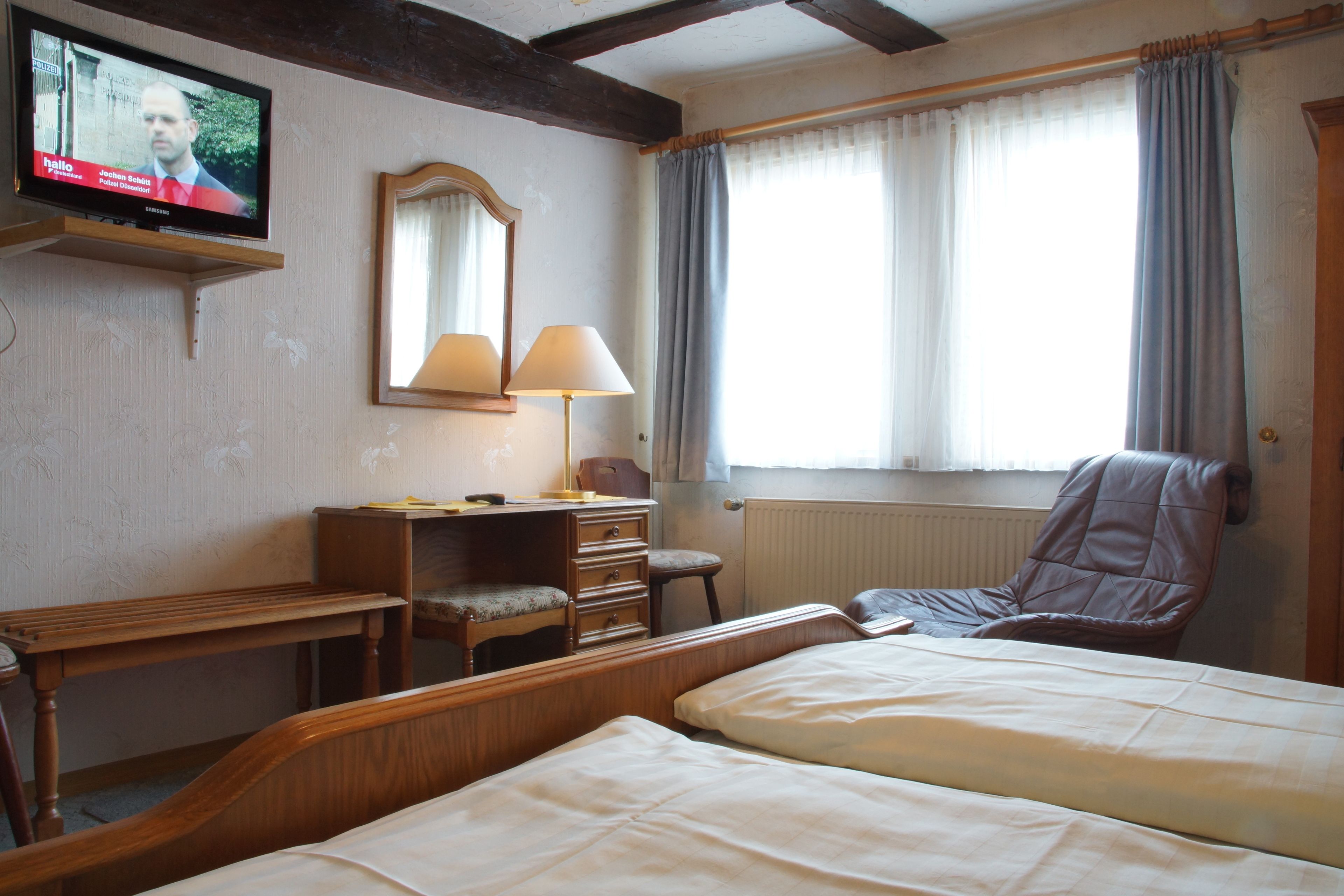 Quarto de hotel com cama, escrivaninha com cadeira, luminária, espelho e televisão na parede.