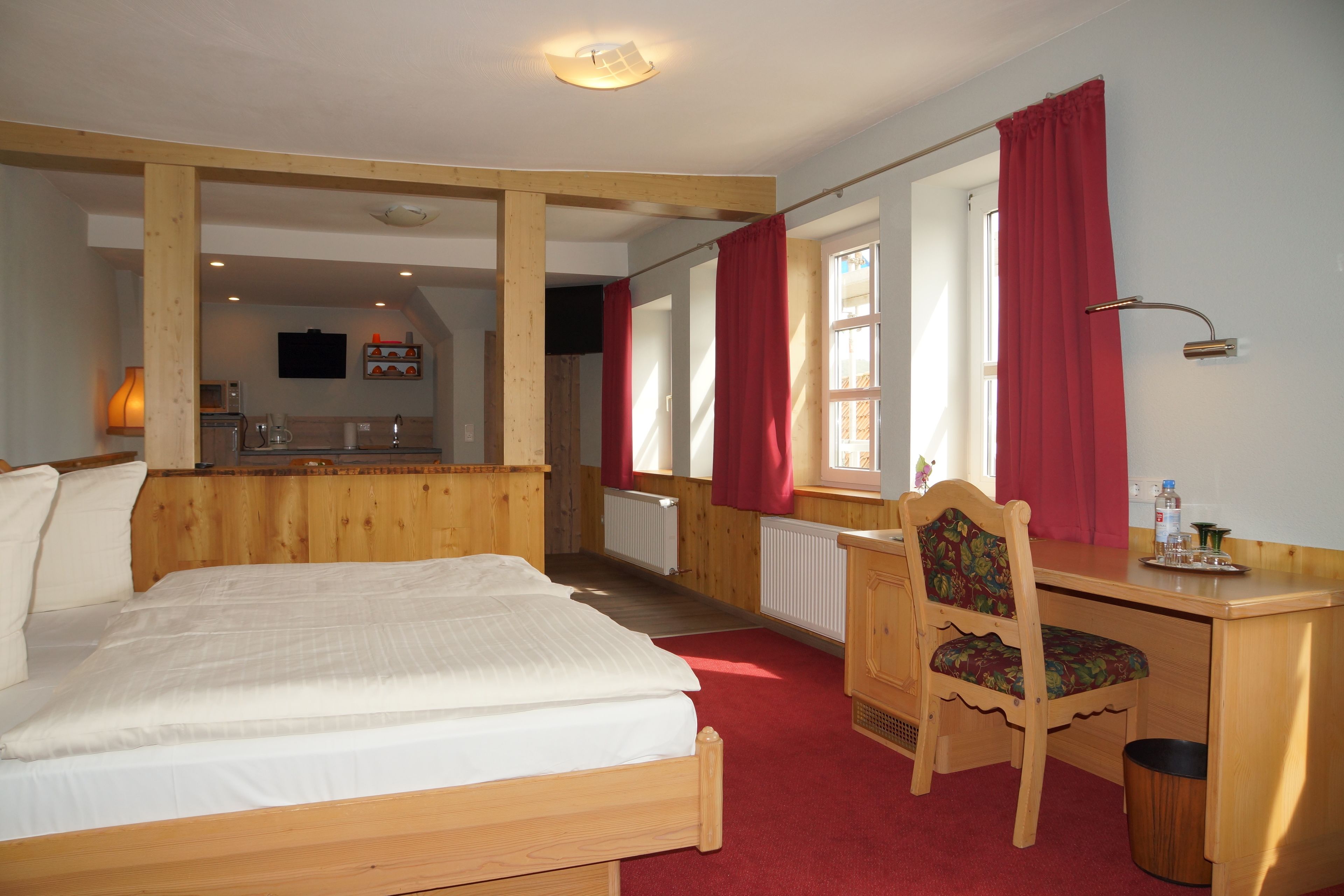 Un dormitorio con cama de madera, un escritorio y una silla. Hay una ventana con cortinas rojas, un radiador y un televisor en la pared.