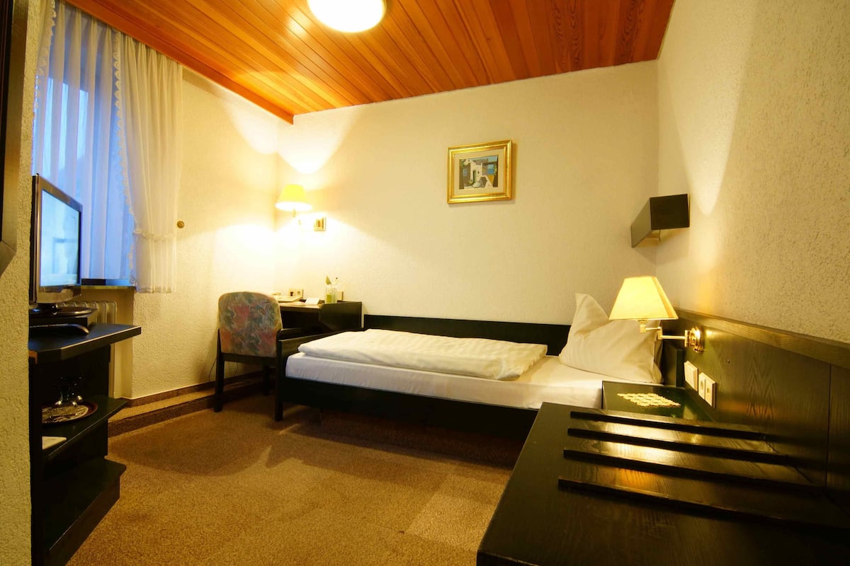 A hotel room with a bed, bedside table, chair, desk, lamp, and a framed picture on the wall.