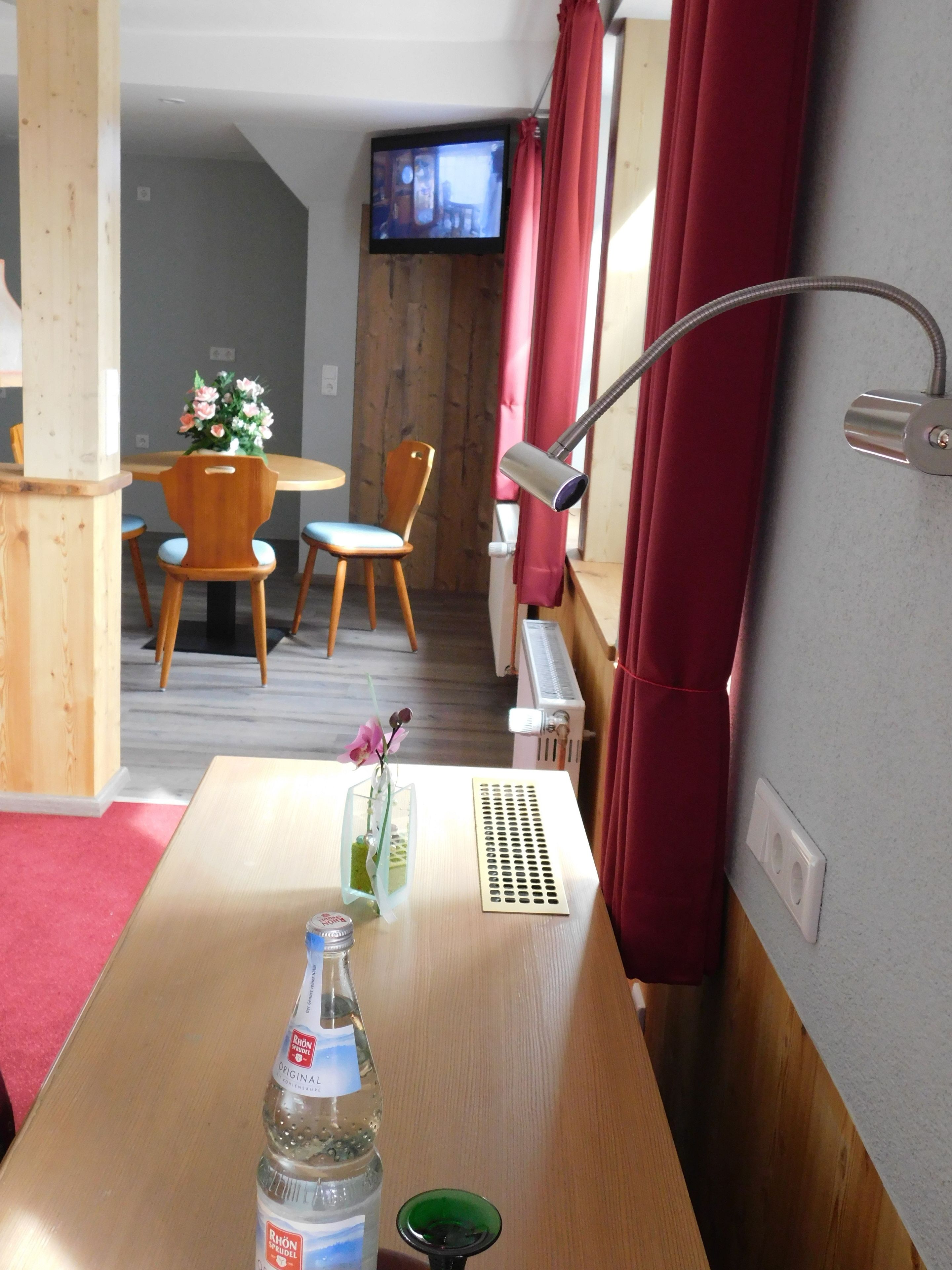 A hotel room with a wooden table, a bottle of water, a green glass, a flower vase, a TV mounted on the wall, and red curtains.