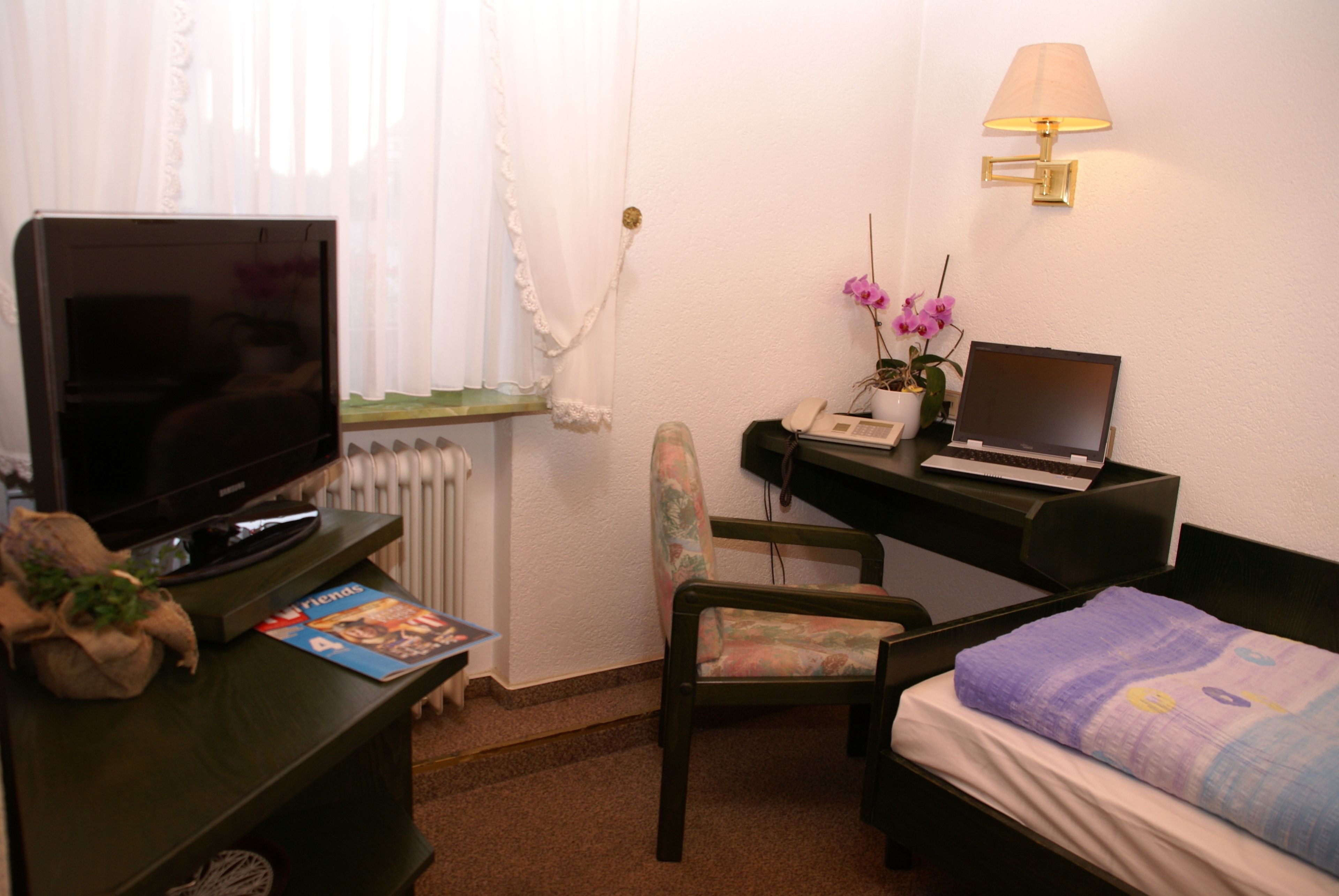 A small, neatly arranged room with a bed, a desk with a laptop, a chair, a television, and a lamp.