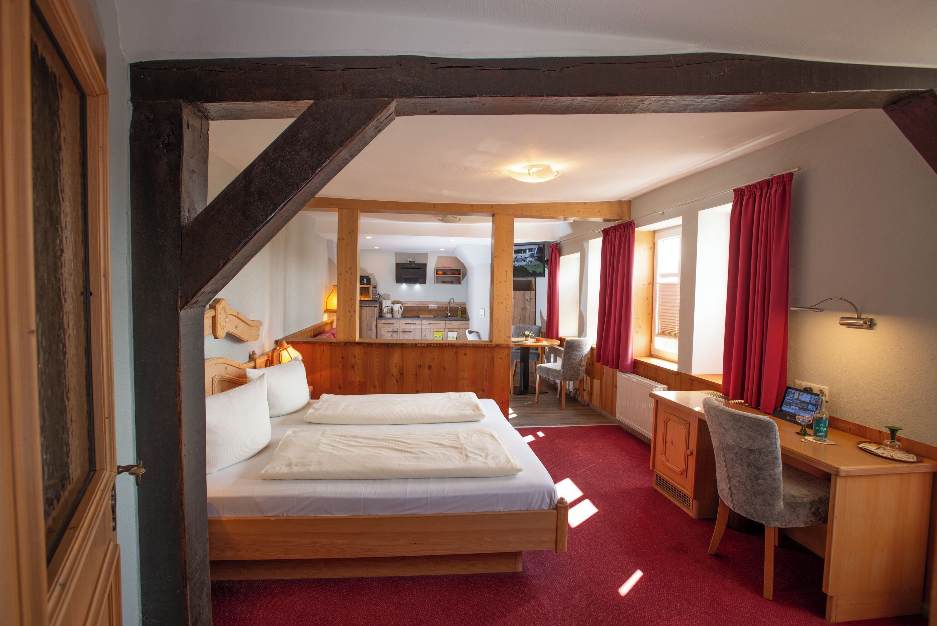 A bedroom with a wooden bed, a desk with a chair, and a view of a kitchen and dining area.