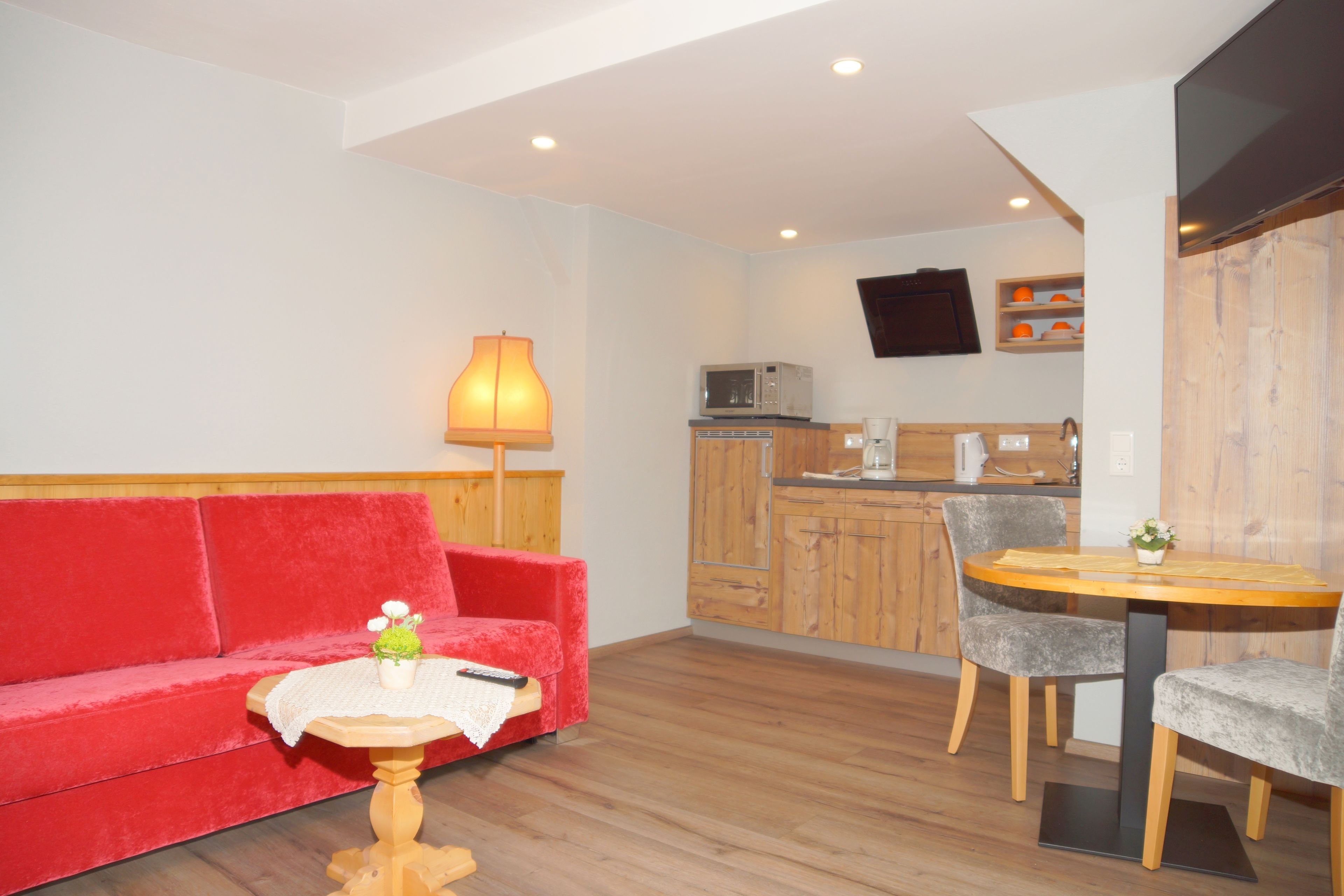 Un espace de vie compact comprenant un canapé rouge, une petite table ronde avec une plante, une table à manger en bois avec deux chaises, un coin cuisine avec un four à micro-ondes et une machine à café, et un téléviseur fixé au mur.