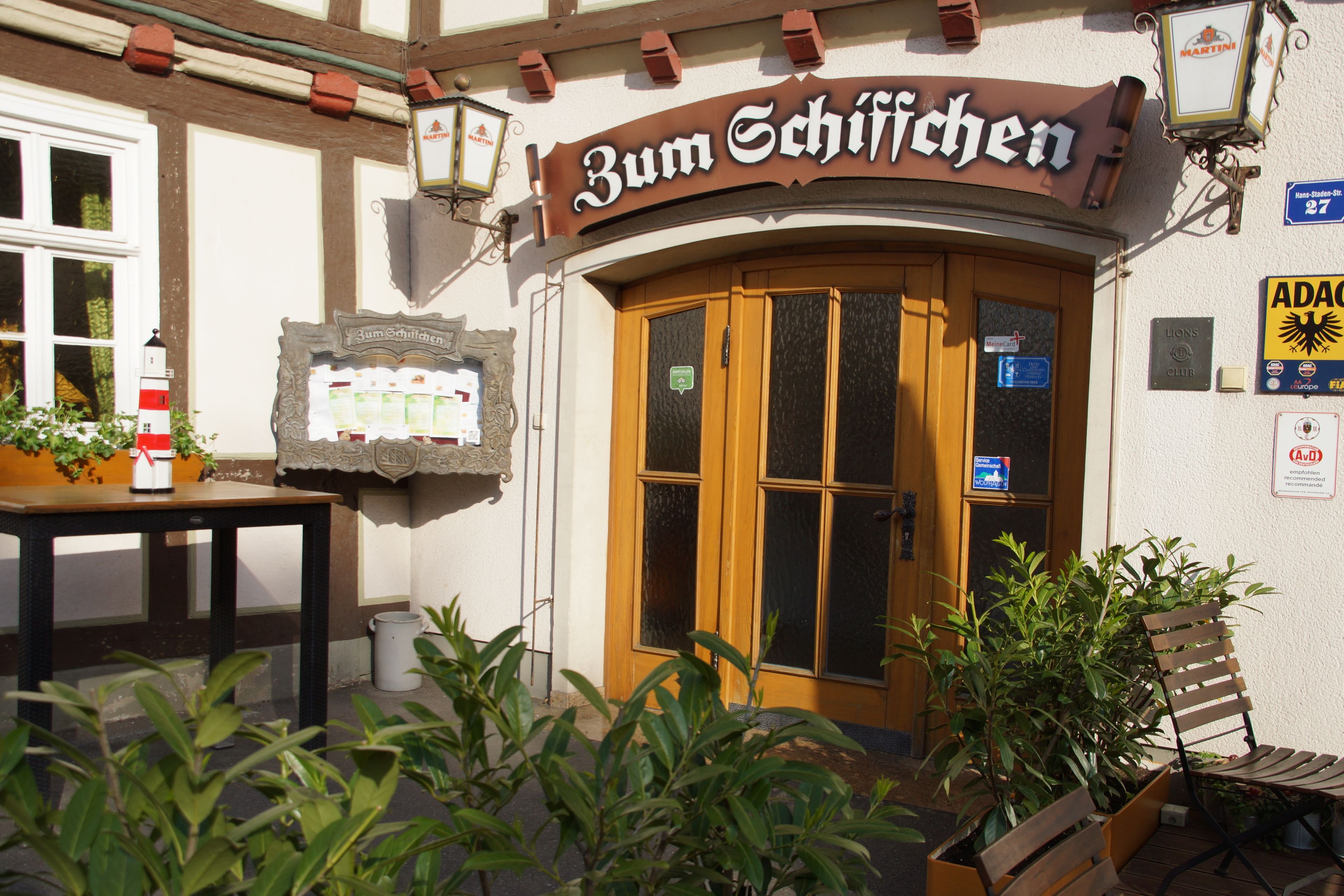 Ein Gebäude mit einem Schild, auf dem "Zum Schifchen" steht, und einer Holztür. Vor dem Gebäude befinden sich Pflanzen und rechts eine Bank.