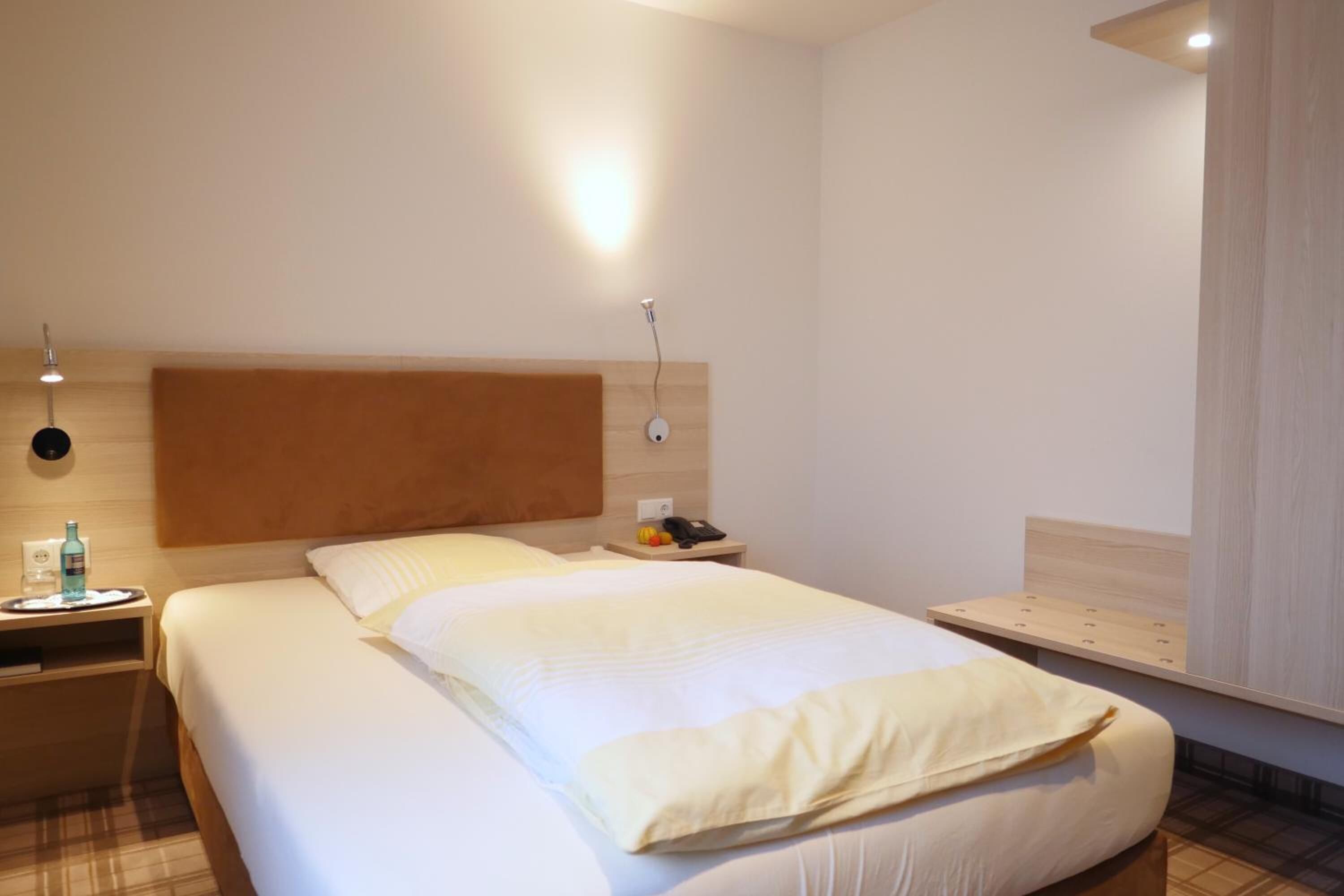 A hotel room with a large bed, a bedside table with a lamp, and a nightstand with a phone and a bottle.