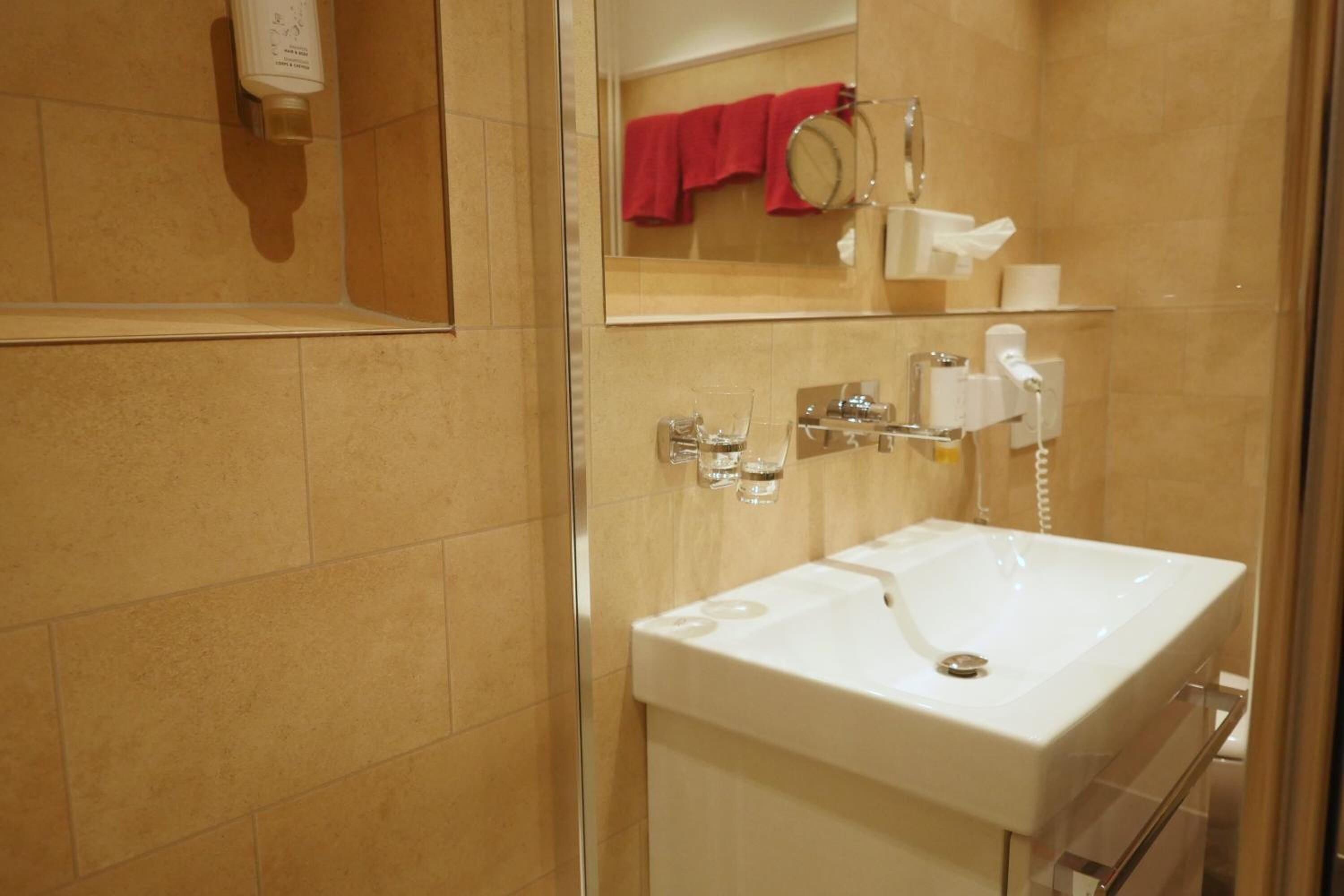 Casa de banho com uma pia branca, um espelho, uma toalha vermelha e cabine de duche em vidro.