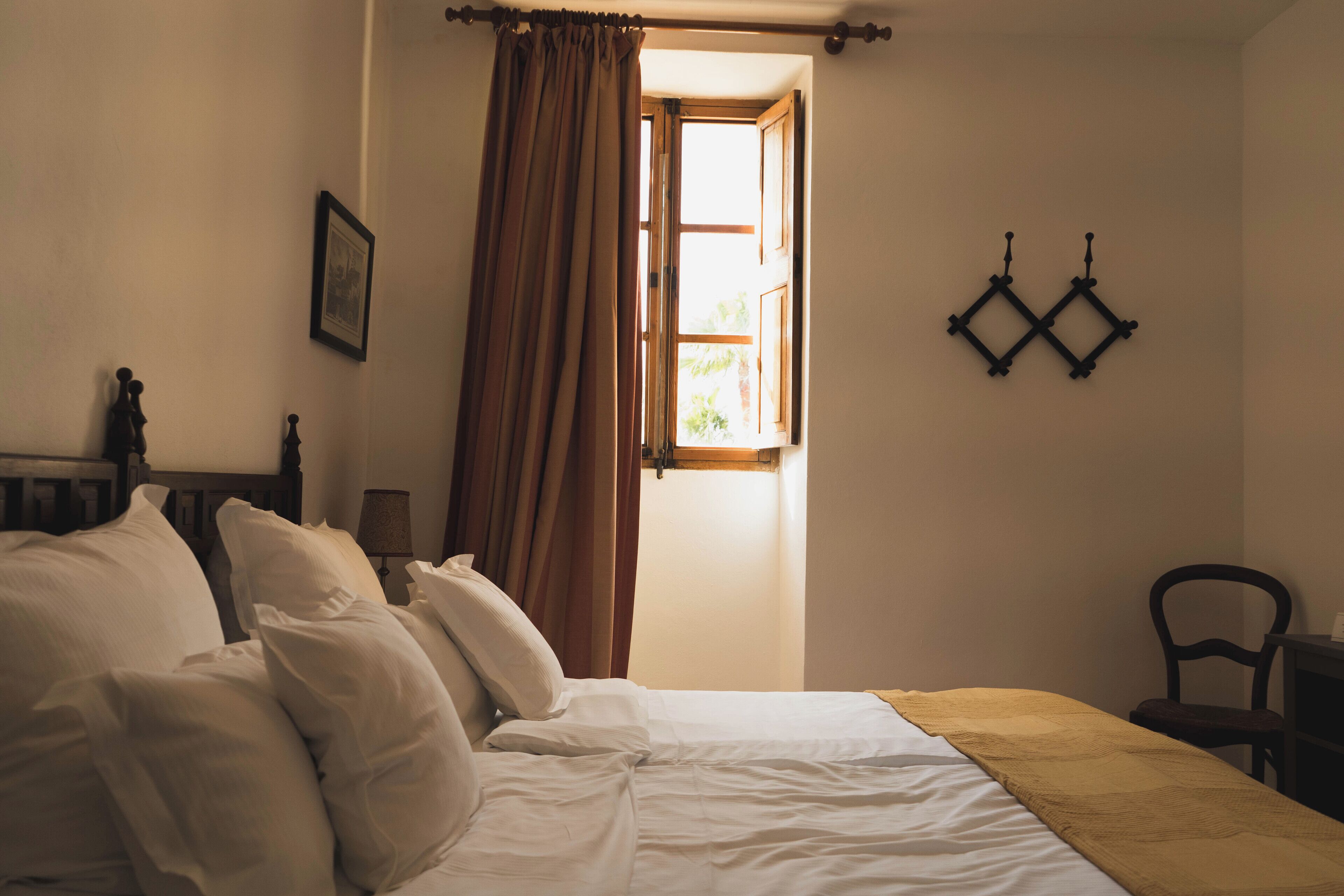 Ein Schlafzimmer mit Bett, einem Fenster mit Vorhängen, einer Wandlampe und einem Sessel.