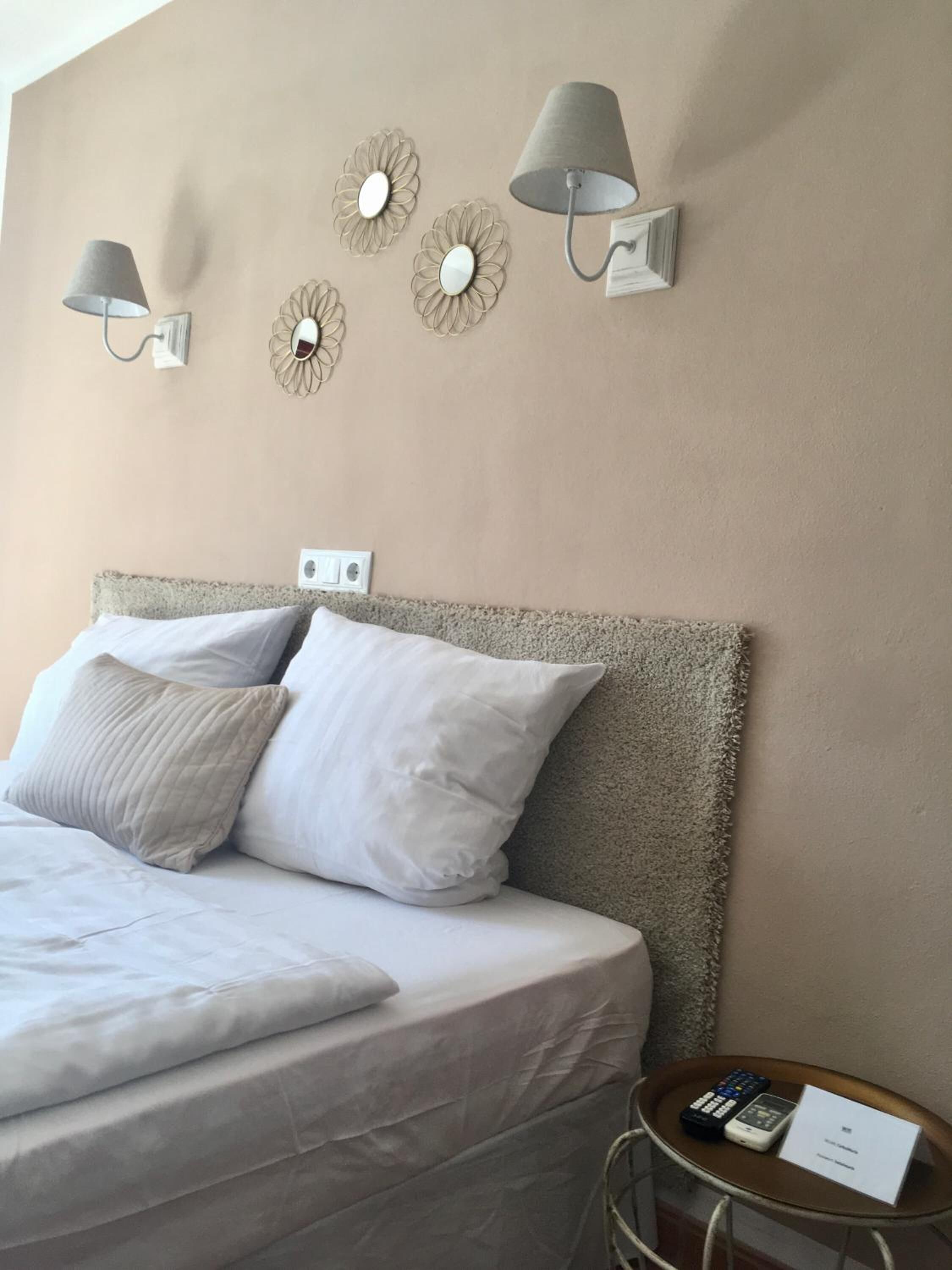 Una cama bien hecha con sábanas blancas y dos almohadas, una mesita con un control remoto y una tarjeta, y lámparas de pared con espejos decorativos.