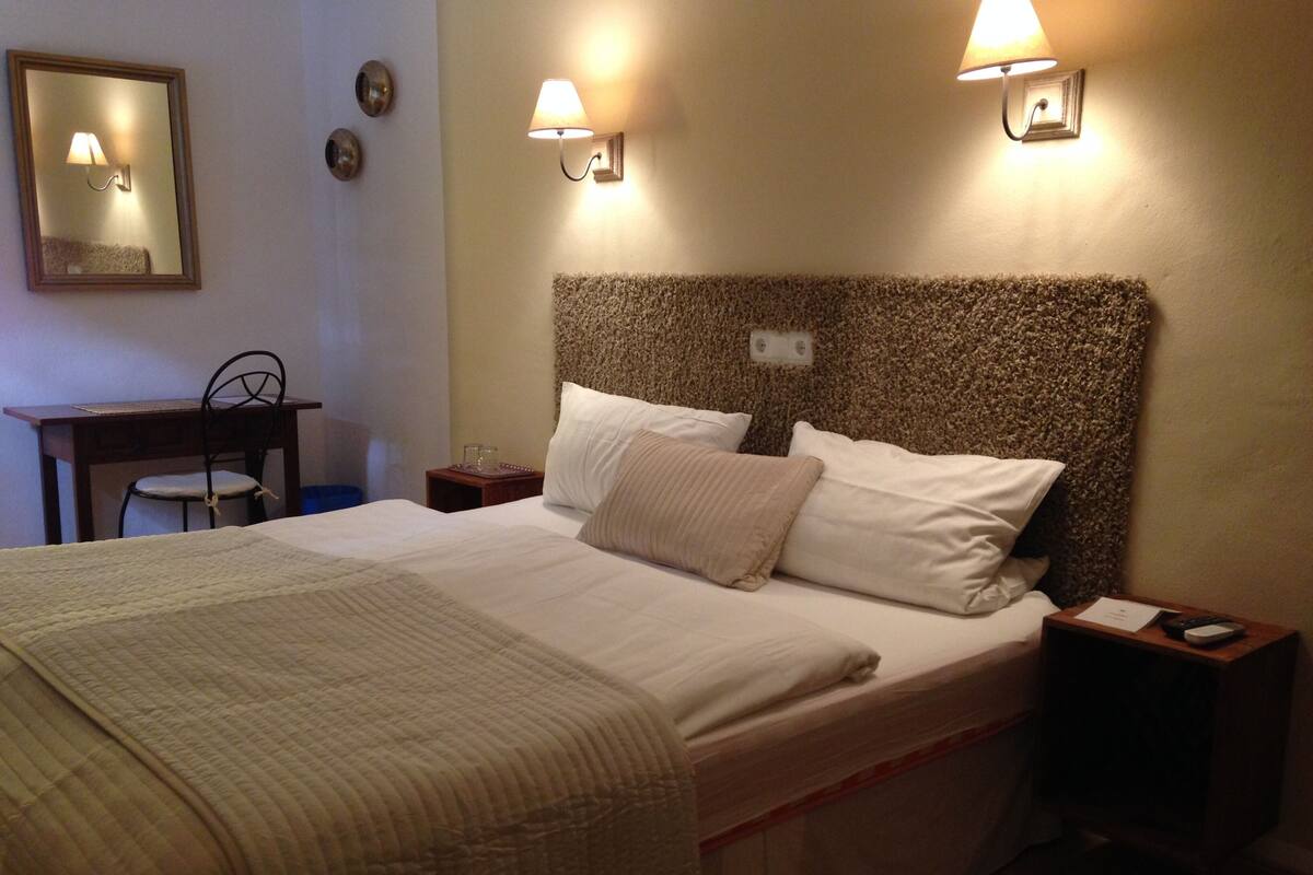 Un lit bien fait, avec une courtepointe, deux tables de chevet, une commode, une chaise et deux lampes fixées au mur.