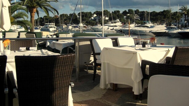 Un espace extérieur aménagé pour les repas, avec des tables et des chaises, donnant sur une marina où sont amarrés des bateaux.
