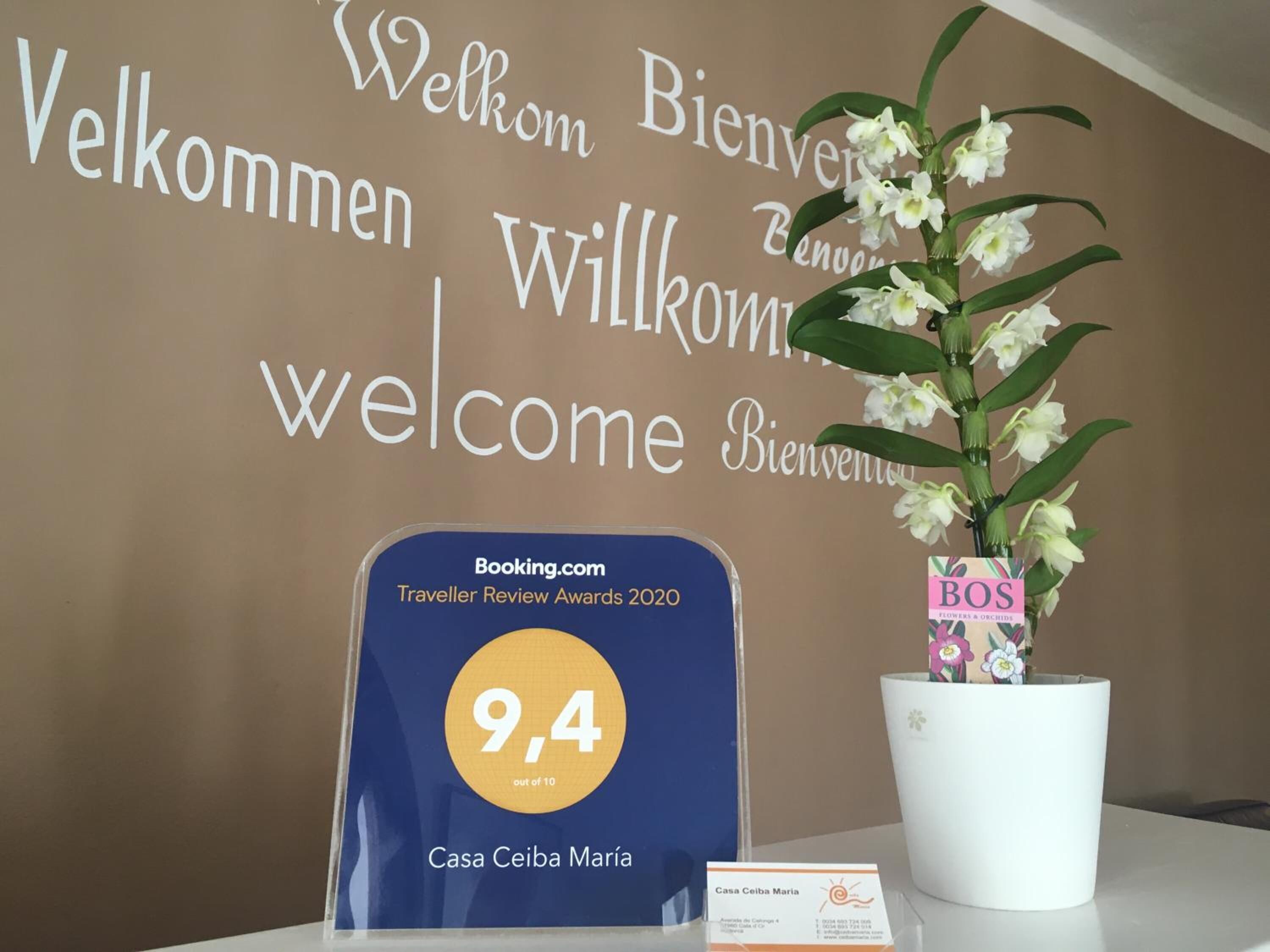 Ein Willkommensschild mit „Welcome“ in verschiedenen Sprachen, eine Auszeichnung von Booking.com und eine Blumenvase.