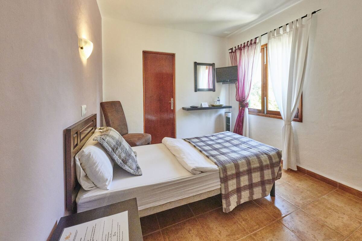 Une chambre à coucher avec un lit, une chaise, un bureau, un miroir et une fenêtre avec des rideaux.