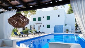 Un espace piscine avec un bâtiment blanc en arrière-plan, un fauteuil suspendu en osier et des chaises longues.