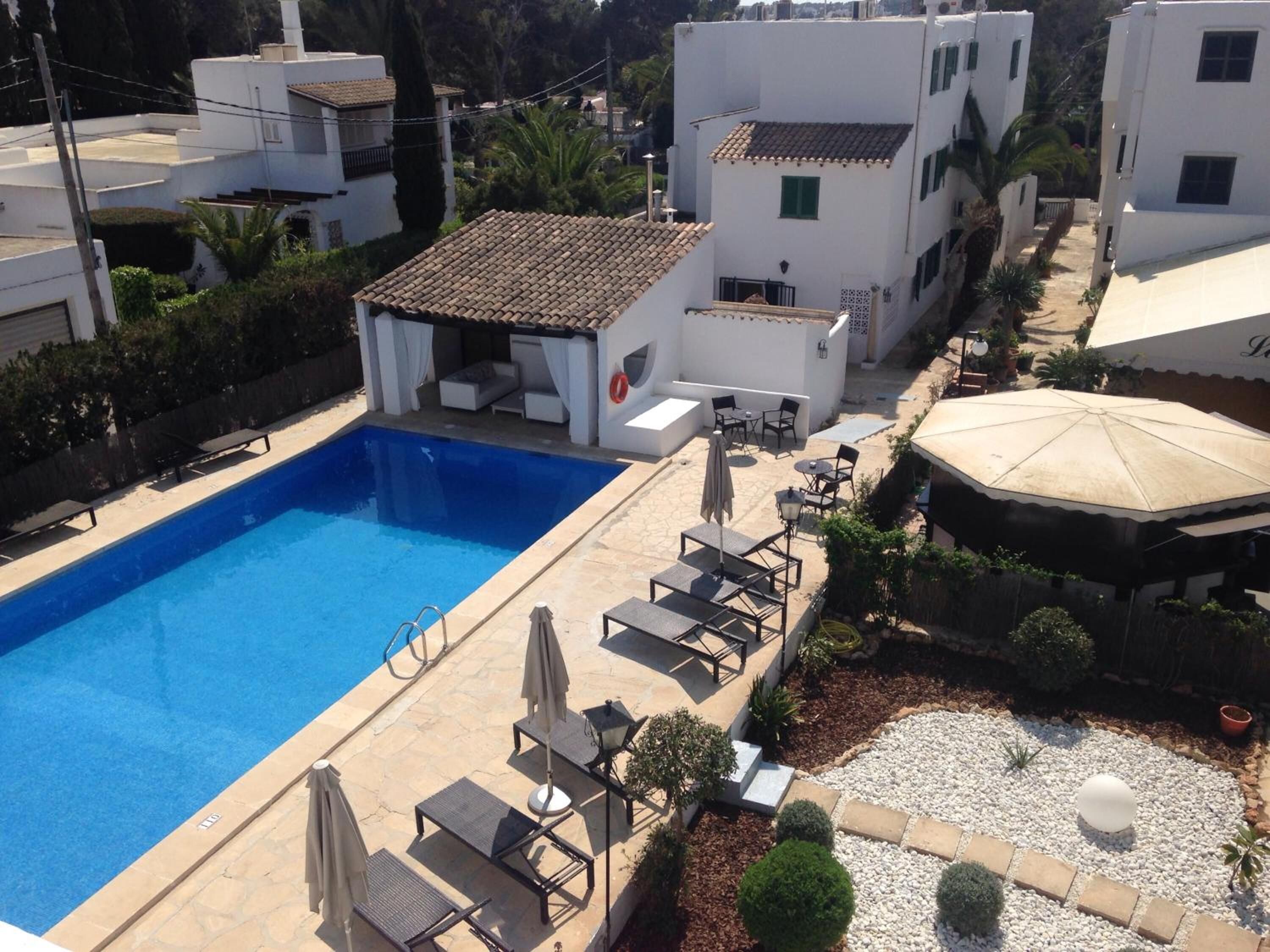 A swimming pool with lounge chairs and umbrellas, surrounded by white buildings and a garden area.