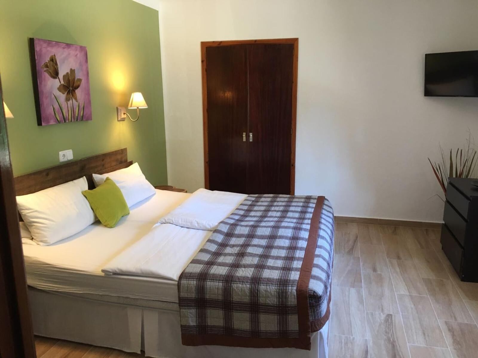 A bedroom with a bed, a wooden wardrobe, a wall-mounted TV, and a painting on the wall.