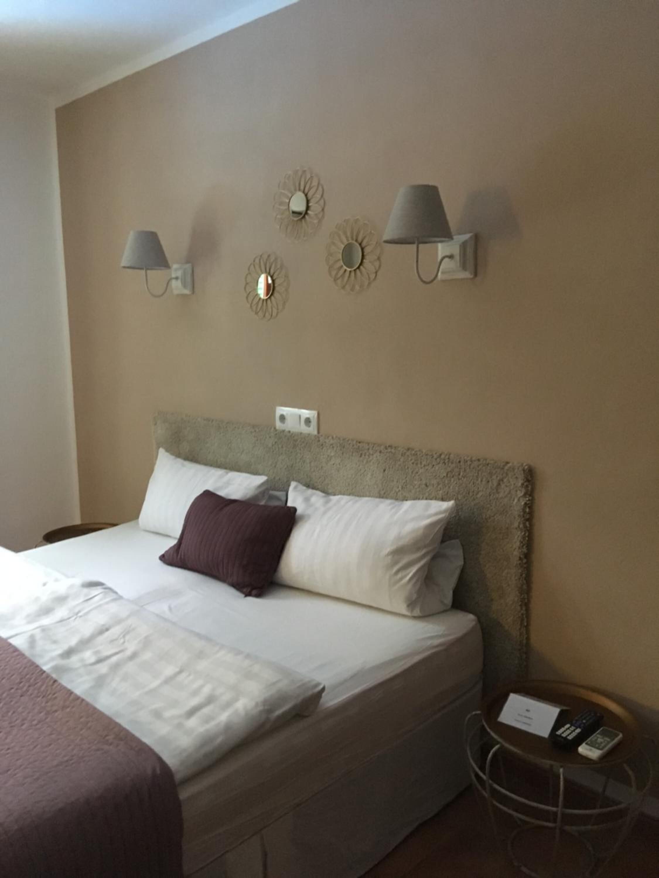 A neatly made bed with white and purple bedding, two wall-mounted lamps, and a small table with a phone and remote control.