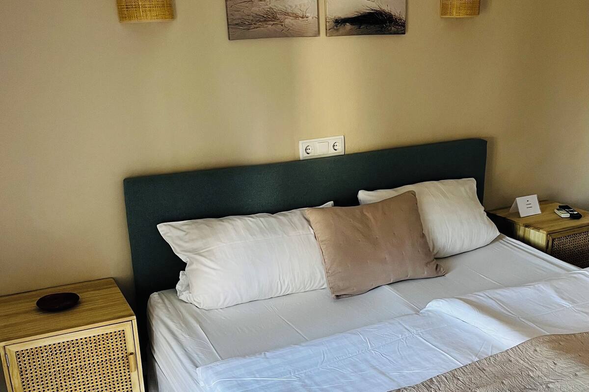 Un lit bien fait, avec du linge de lit blanc et beige, une tête de lit vert foncé et deux lampes fixées au mur.