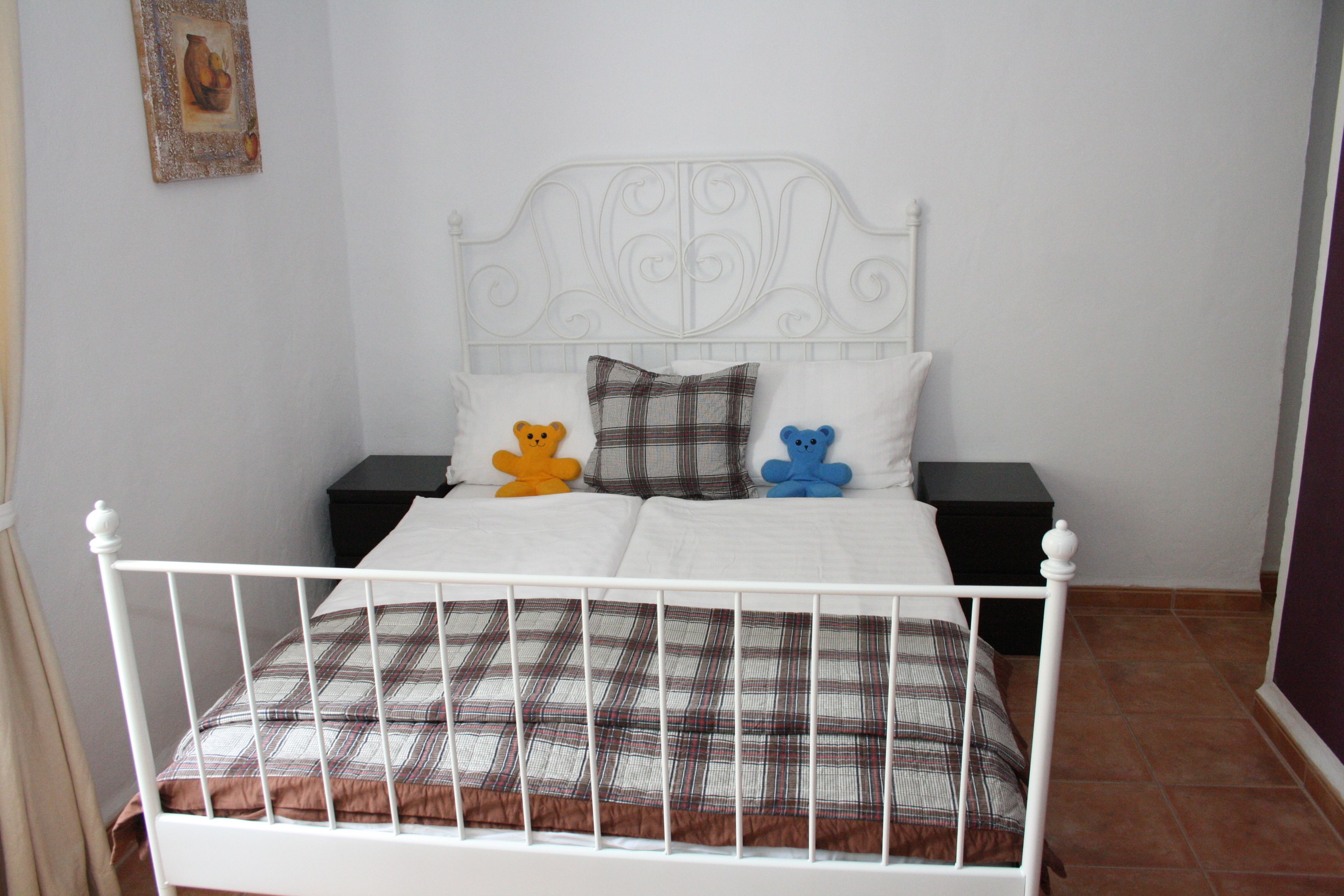 Una cama de metal blanco con una colcha escocesa y dos almohadas, una con un diseño de oso y otra con estampado de cuadros.