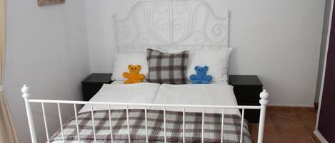 Una cama de metal blanco con una colcha escocesa y dos almohadas, una con un diseño de oso y otra con estampado de cuadros.