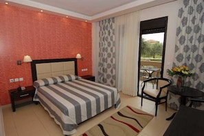 A hotel room with a bed, bedside tables, a chair, a small table with flowers, and a view through a door. - Astir Notos (Thasos)