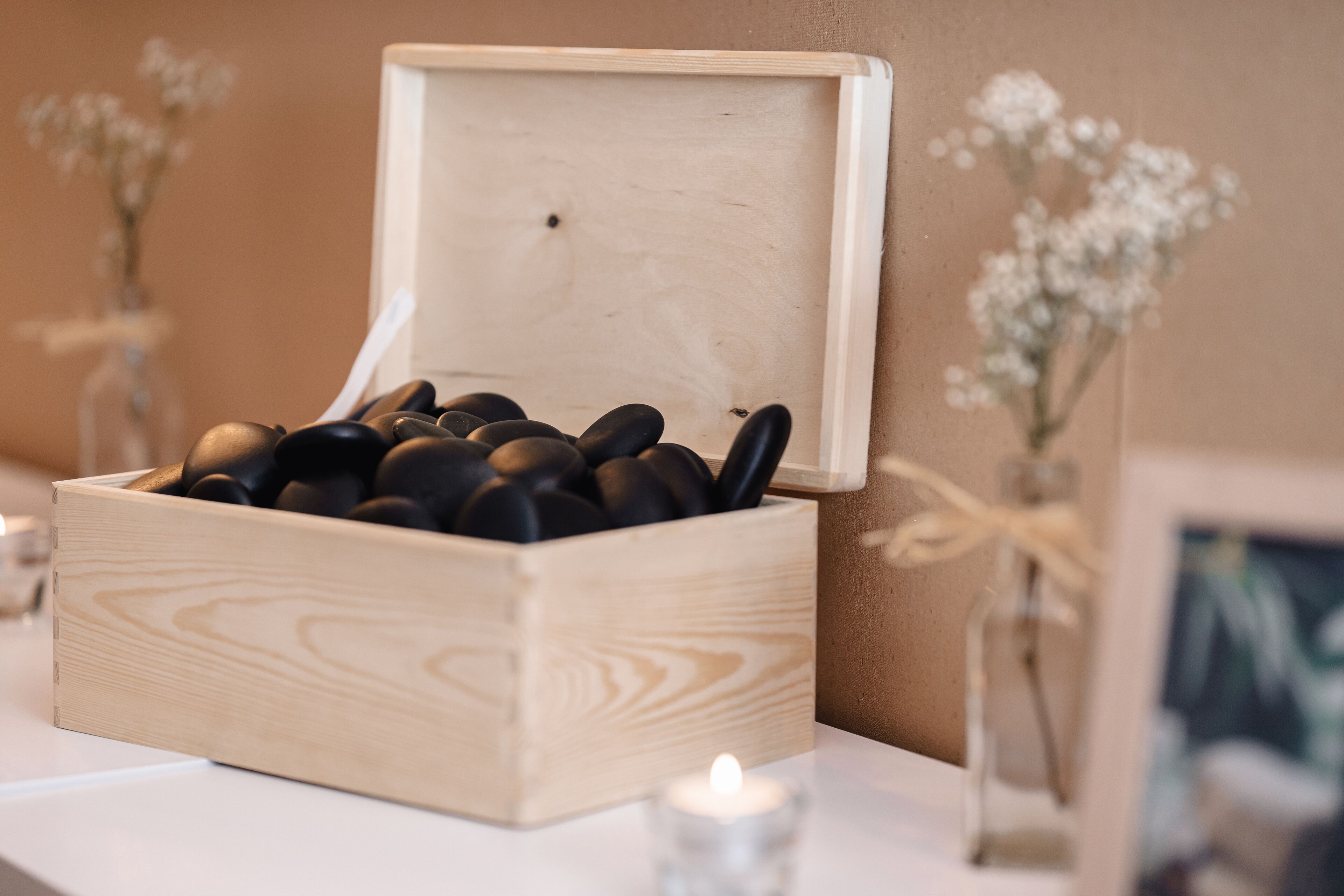 Een houten doos gevuld met zwarte stenen op een witte ondergrond, met een brandende kaars ervoor.