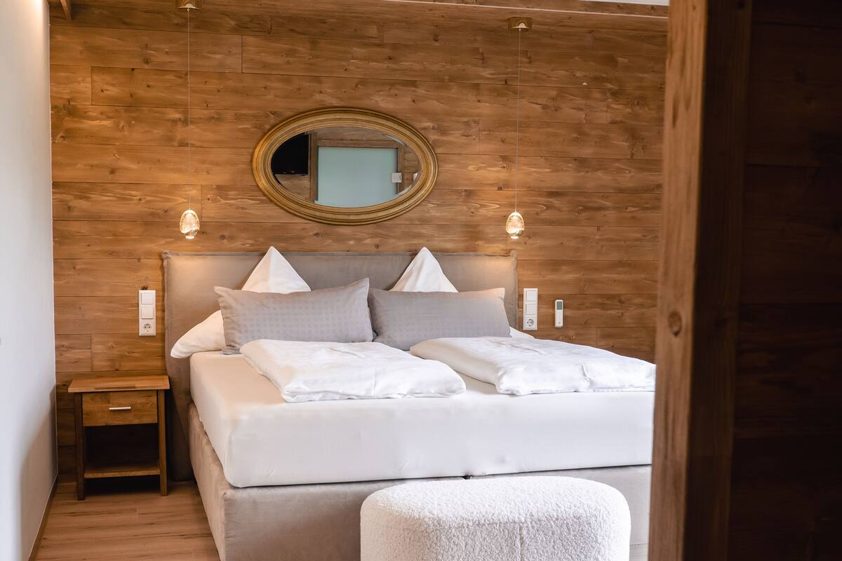 A bedroom with a wooden headboard, a round mirror, a bedside table, and a white ottoman.