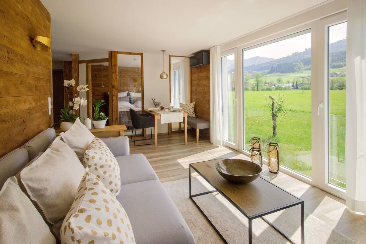 A modern living room with a sofa, coffee table, and a view of a dining area and outdoor landscape.