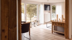 Una habitación moderna con suelo de madera, una mesa de comedor con sillas, una consola de madera con velas y ventanas grandes con vistas a árboles y una valla.