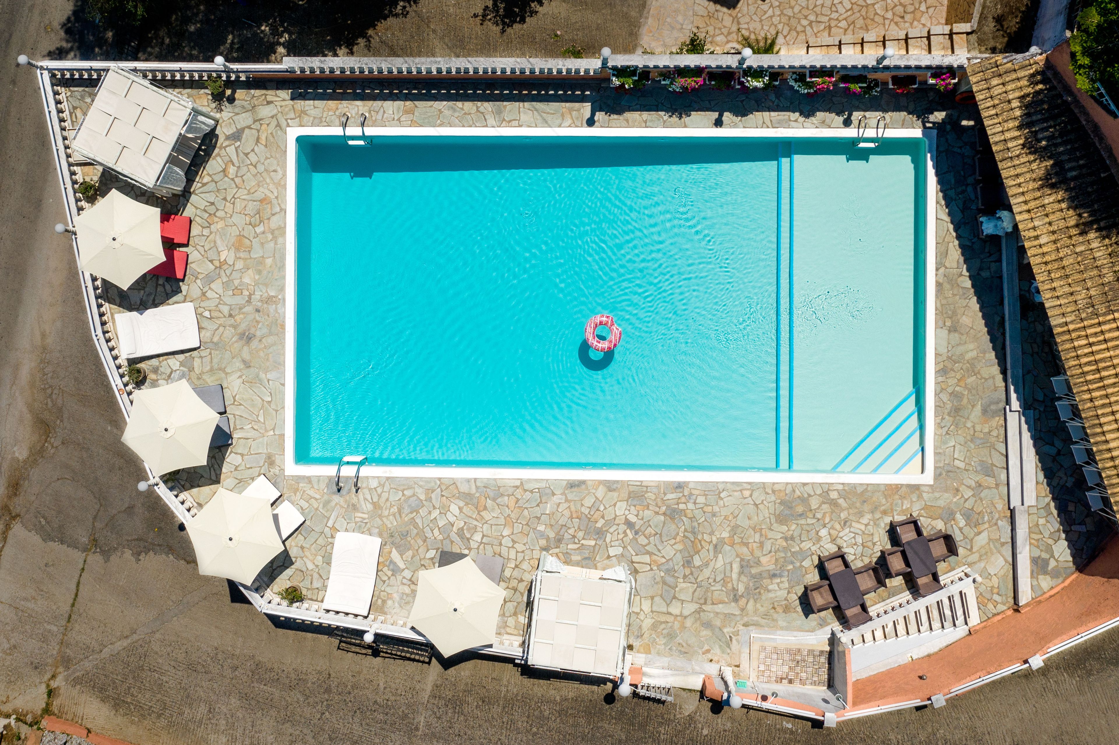 Une vue aérienne d’une piscine rectangulaire avec un seul flotteur gonflable, entourée de chaises longues et de parasols, dotée d’une terrasse carrelée en pierre et d’un mur en pierre.