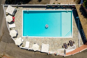 An aerial view of a rectangular swimming pool with a single inflatable ring, surrounded by lounge chairs and umbrellas, with a stone-tiled deck and a stone wall. - Victoria Hill (Corfu)