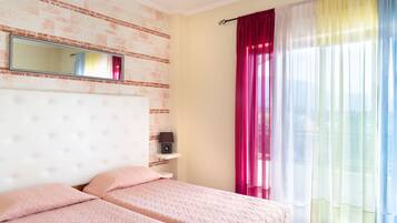 A bedroom with a brick-patterned wall, a white tufted headboard, two beds with pink bedding, a bedside table with a lamp, and a window with sheer curtains.
