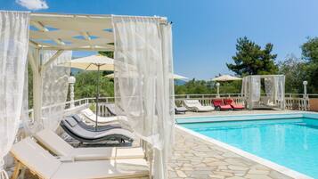 A poolside area with lounge chairs, a pool, and a pergola with white curtains.