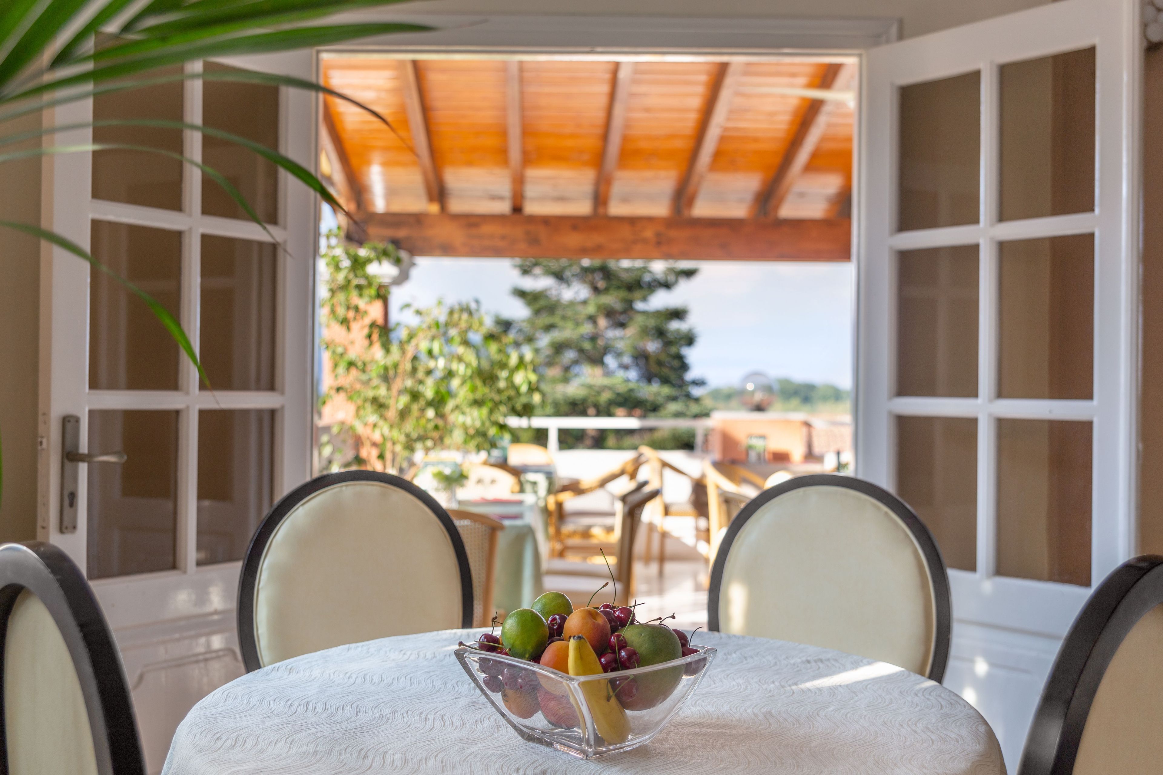 A table with a fruit bowl, two chairs, and a view of a garden through open doors.