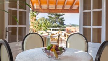 A table with a fruit bowl, two chairs, and a view of a garden through open doors.