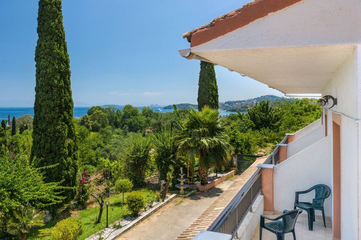A balcony with a view of the sea, surrounded by greenery and tall trees.