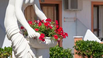 A white statue of a woman holding a flower pot with red flowers, set on a pedestal surrounded by a variety of colorful flowers.
