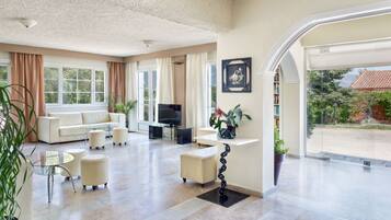 A spacious living room with a sofa set, a TV, and a coffee table. There are potted plants and a bookshelf.