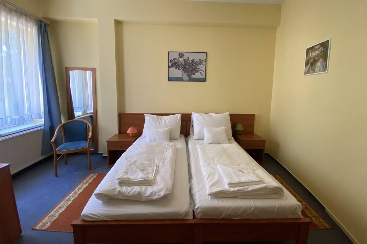 A hotel room with two single beds, a desk, a chair, a window with curtains, and two paintings on the walls.