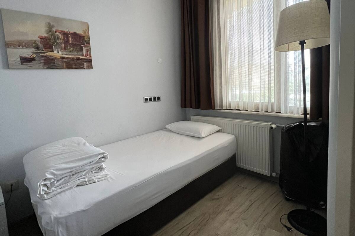 Ein schlichter, sauberer Raum mit einem Einzelbett, einem Heizkörper, einer Lampe und einem Bild an der Wand.
