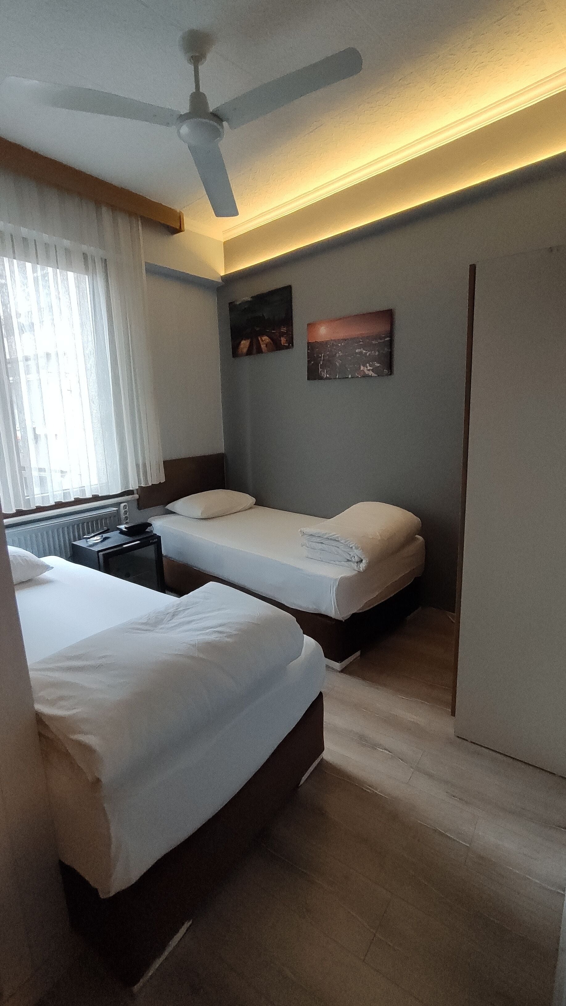 Habitación de hotel con dos camas, ventilador de techo, radiador y dos cuadros en la pared.