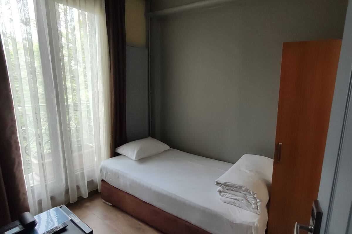 Ein kleines Zimmer mit einem Einzelbett, einer Holztür, einem Nachttisch und einem Fenster mit durchsichtigen Vorhängen.