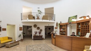 A spacious lobby with a high ceiling, a reception desk, a yellow sofa, a staircase leading to a balcony, and various decorative items. - Hotel Su Baione (Abbasanta)