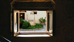 Une vue à travers une fenêtre à arc, dévoilant un petit jardin avec une fontaine, des plantes en pot et une structure en pierre.
