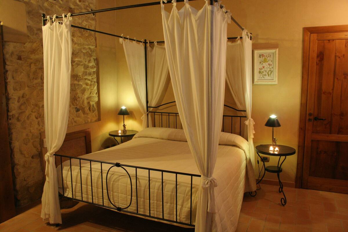 A four-poster bed with white curtains, a stone wall, a wooden door, and two bedside tables with lamps.