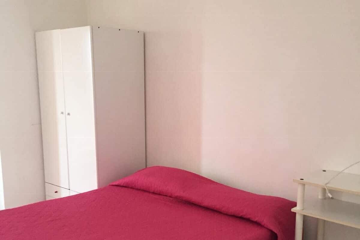 A small, single-bed room with a red bedspread, a white wardrobe, and a white shelf unit.