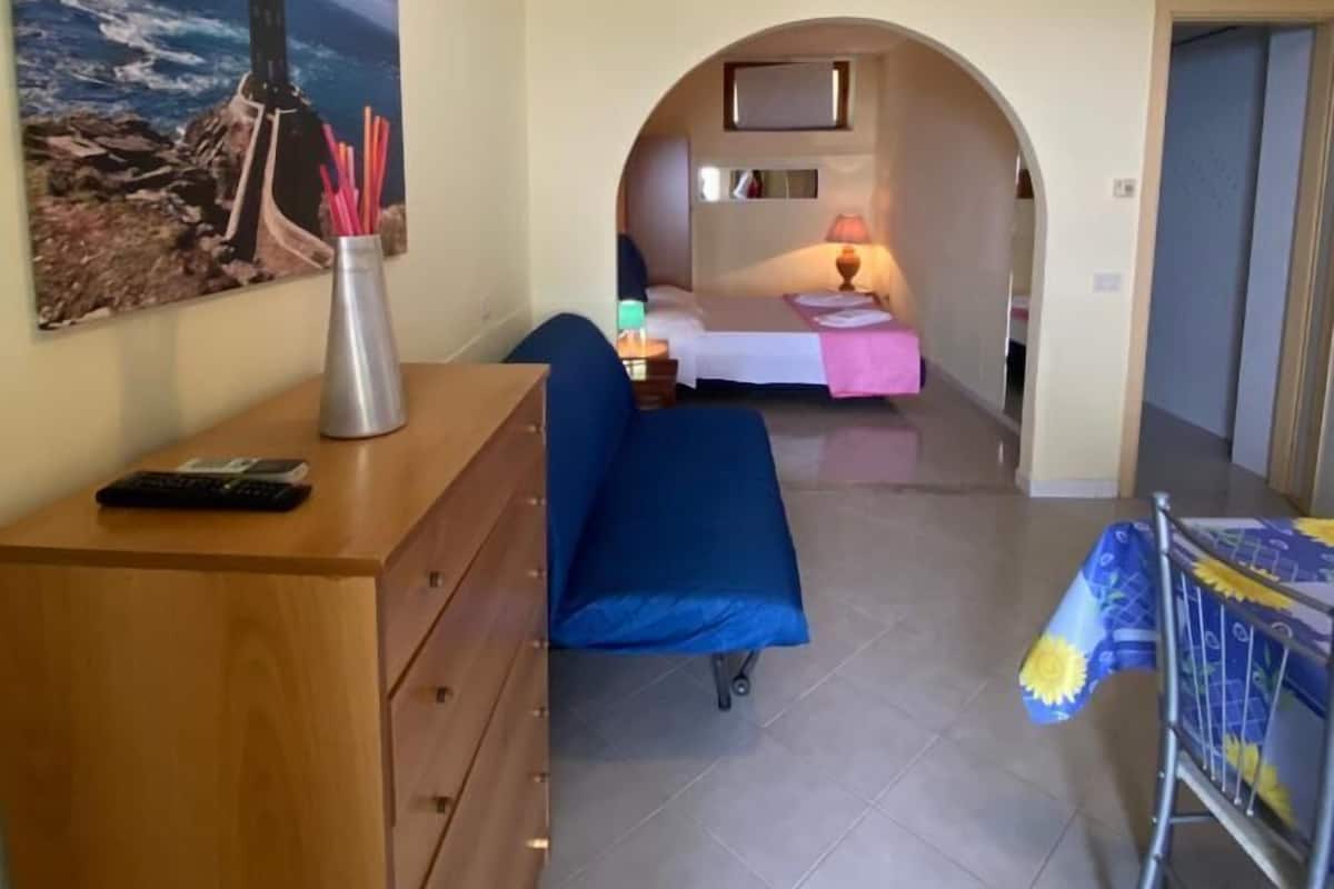 A room with a wooden dresser, a bed with a pink blanket, and a painting of a lighthouse.
