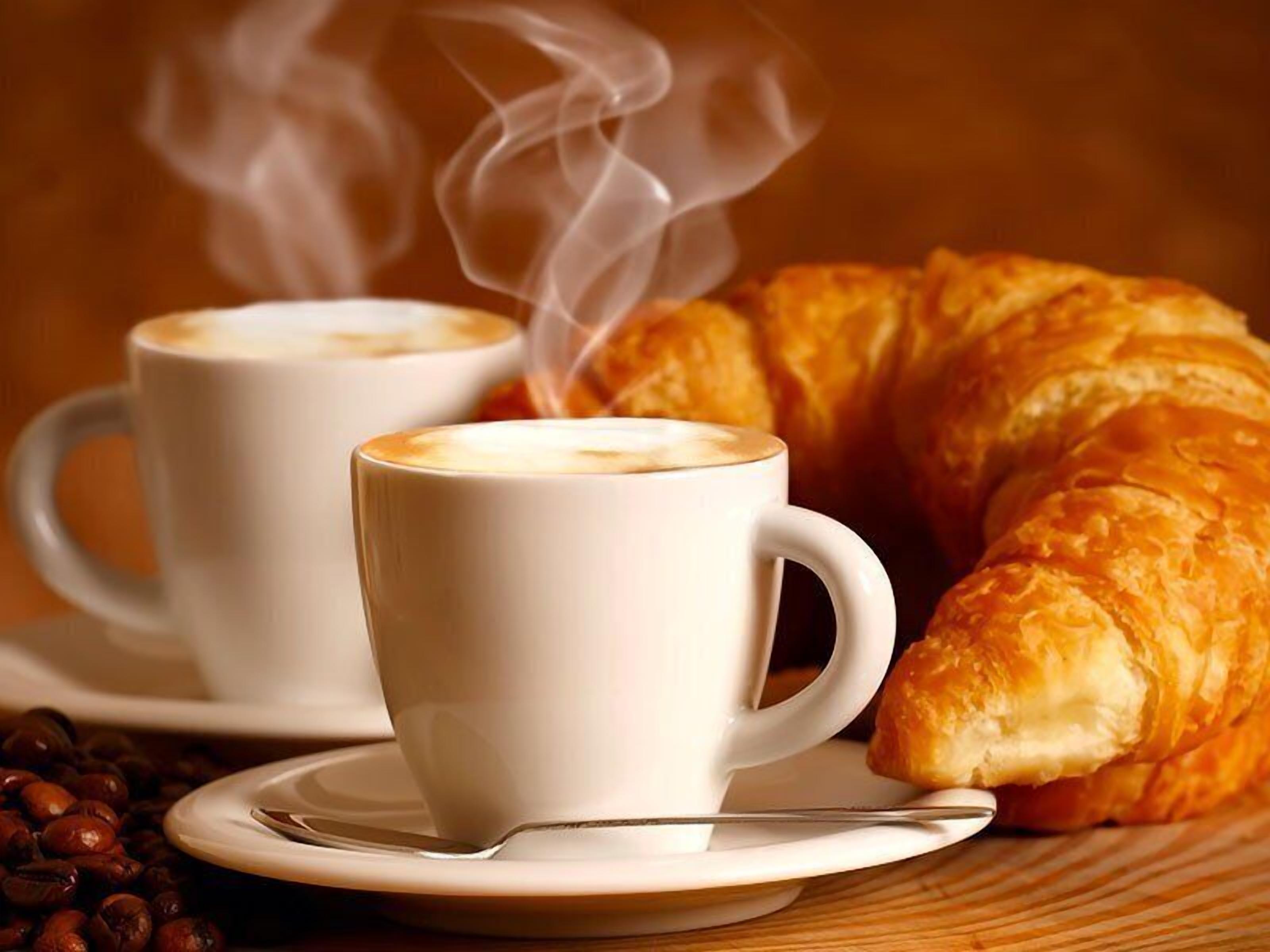 Two white cups of coffee with steam, a croissant, and coffee beans on a wooden surface.