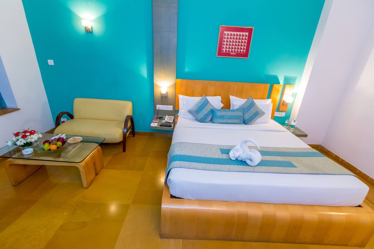 A hotel room with a bed, a sofa, a glass coffee table with fruit, and a wall clock.