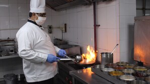 A chef in a white uniform with a hat, handling a pan with flames on a stove in a kitchen. - Capitol Hill (Ranchi)