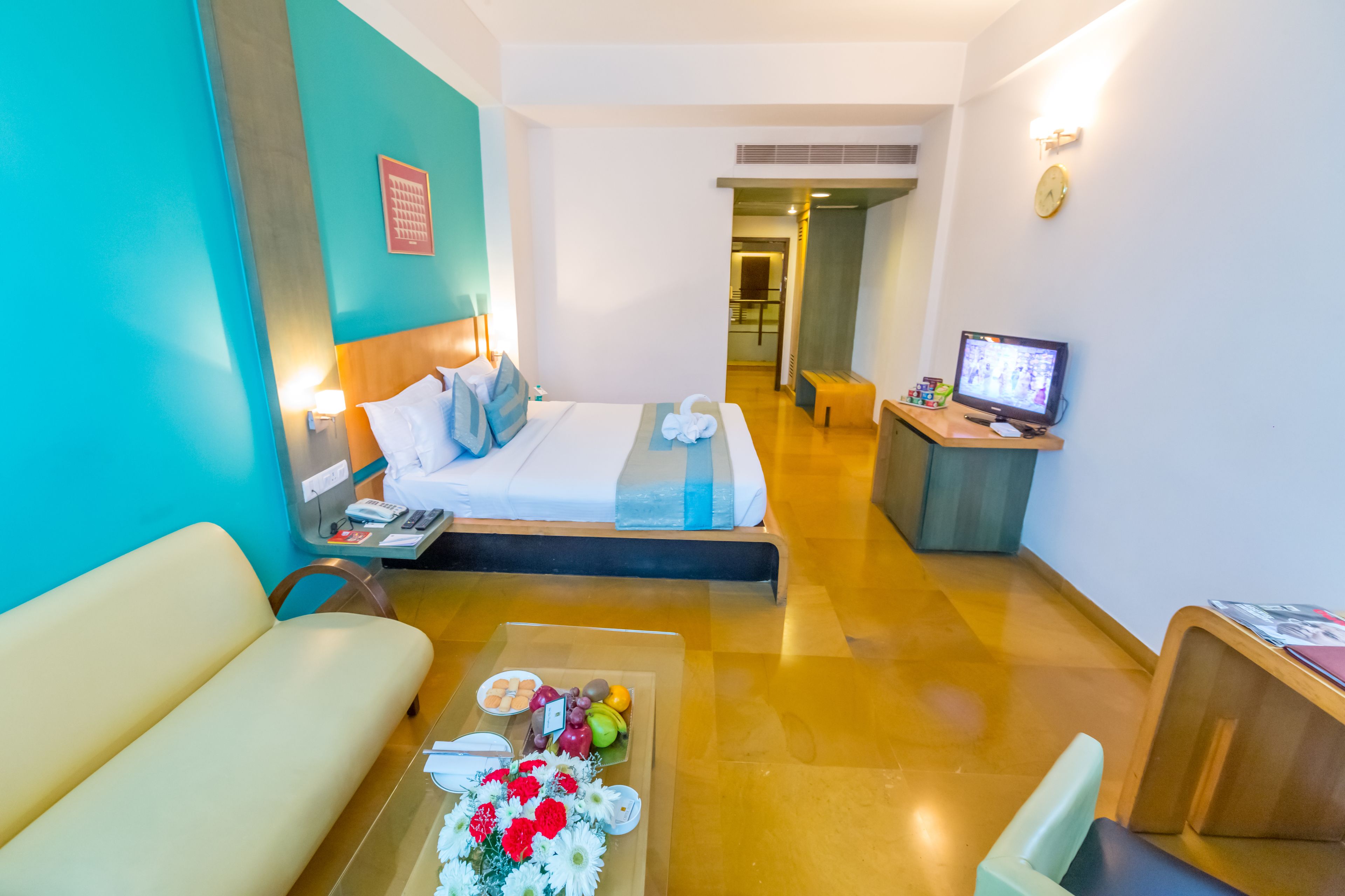 A hotel room with a bed, a desk with a computer, a sofa, and a coffee table with flowers and fruits.